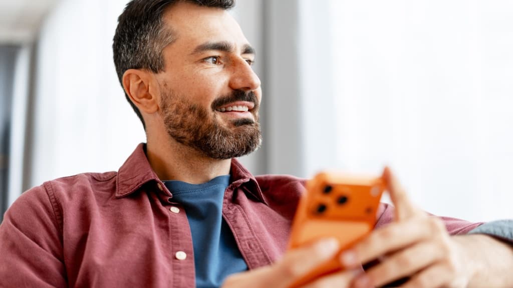 Smiling bearded man smiling and looking away while holding a modern smartphone.
