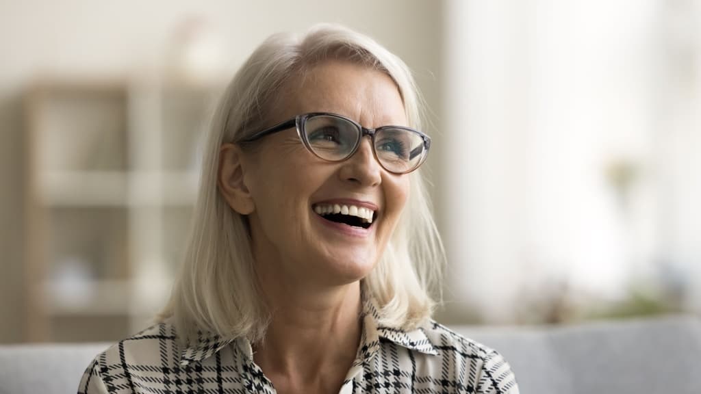 Happy beautiful blonde mature woman in elegant glasses sitting on couch