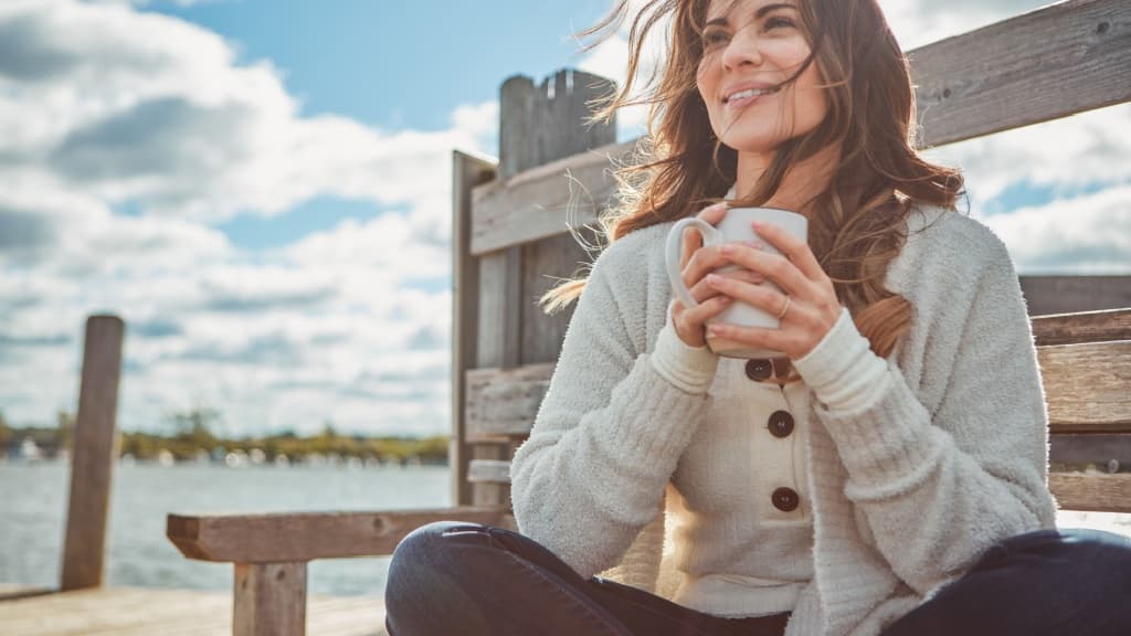 Woman sat by a lake drinking a coffee.