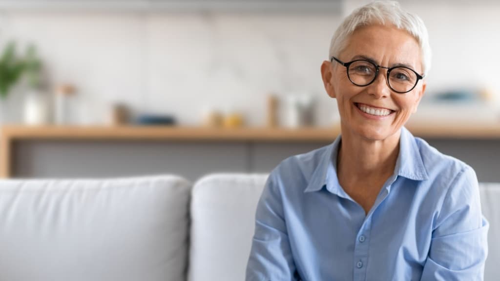 A cheerful older woman with gray hair and glasses beams joyfully while sitting comfortably on a soft couch